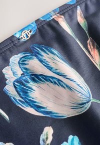 Tela oscura con un gran estampado floral de tulipanes en azul y blanco y un pequeño logo metálico dorado redondo cerca del borde cosido.