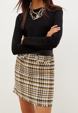 Kobieta ze skrzyżowanymi ramionami, ubrana w czarną bluzkę z długim rękawem, masywny złoty i przezroczysty łańcuchowy naszyjnik oraz mini spódniczkę w pepitkę w kolorach żółtym, czarnym i białym, z przednimi kieszeniami.