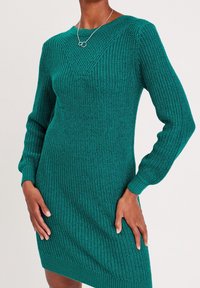 Robe en tricot de couleur bleu sarcelle vif avec un motif texturé. Comprend des manches longues, des poignets côtelés et un décolleté en V. Coupe mi-longue.