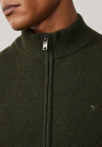 Olijfgroene zip-up trui met geribde hoge kraag en textuurgebreide stof. Bevat een klein logo boven de rits. Zilveren hardware.