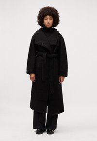 Cappotto lungo nero e fluffy con ampio colletto, cintura a cravatta e grandi tasche frontali. Presenta un tessuto testurizzato e maniche larghi.