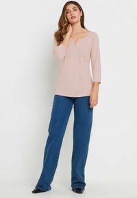 Haut rose clair à manches longues avec un col rond et trois boutons ; associé à un jean droit bleu foncé et des chaussures noires à bout pointu.