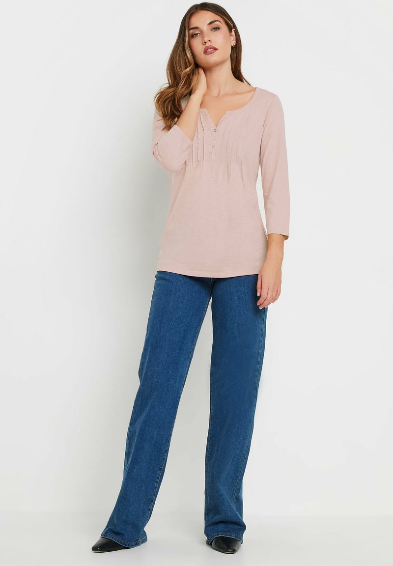 Haut rose clair à manches longues avec un col rond et trois boutons ; associé à un jean droit bleu foncé et des chaussures noires à bout pointu.