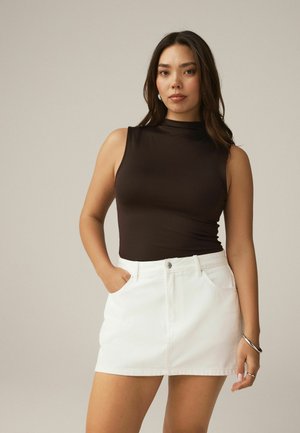 Kvinde med langt mørkt hår iført sort ærmeløs top og hvid denimskørt, stående med den ene hånd i lommen mod en ensfarvet baggrund.