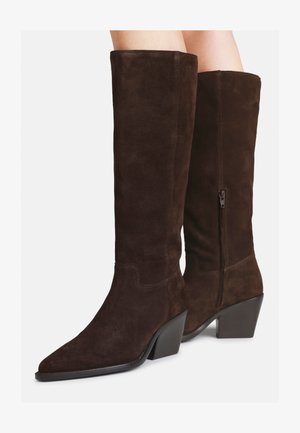 Bottes en daim marron foncé, montantes jusqu'au genou, avec des bouts pointus, des talons bloc et des fermetures éclair latérales, portées sur des jambes nues sur fond blanc.