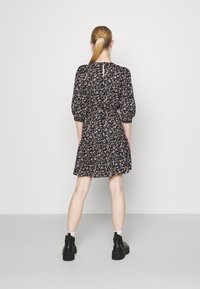 Vrouw met een blonde paardenstaart, gekleed in een zwart bloemenjurk, witte sokken en zwarte enkellaarsjes, staat met haar gezicht naar een eenvoudige witte muur.