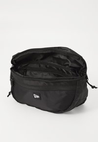 Sac en nylon noir de forme arrondie, avec des compartiments zippés doubles et des sangles réglables. Présente un petit logo à l'avant.
