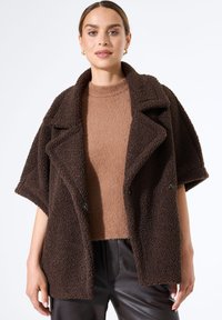 Braune strukturierte Außenschicht mit einem breiten Schalkragen und kurzen Ärmeln, versehen mit schwarzen Knöpfen, über einem hellbraunen Stricktop.