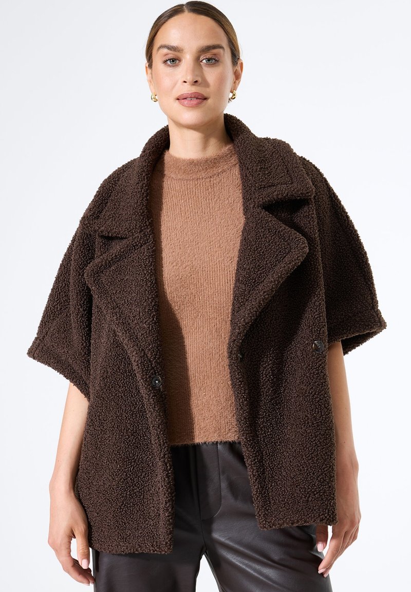 Braune strukturierte Außenschicht mit einem breiten Schalkragen und kurzen Ärmeln, versehen mit schwarzen Knöpfen, über einem hellbraunen Stricktop.