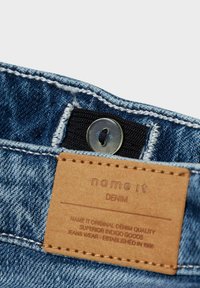 Denimtyg i en medelblå nyans, med en läderlapp märkt "name it" och en rund knapp ovanför ett svart midjeband.