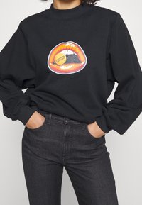 Sudadera negra con un gráfico de labios naranja brillante y un logotipo redondo amarillo de "Wrangler", de corte relajado con puños y cuello acanalados.