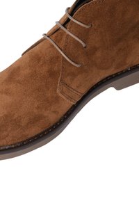 Stivale alla caviglia in suede marrone con una texture liscia, punta rotonda e lacci beige. La suola è scura con cuciture visibili.