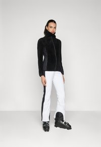 Jachetă moltonată neagră cu guler înalt și fermoar în față, combinată cu pantaloni albi cu dungi negre pe laterale și bocanci de schi. Textură de țesătură netedă.
