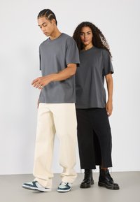 To personer iført løst siddende crewneck t-shirts i mørkegrå. Ét par cremefarvede, vide bukser og en sort, knælang nederdel. Fodtøj inkluderer sneakers og sorte støvler.