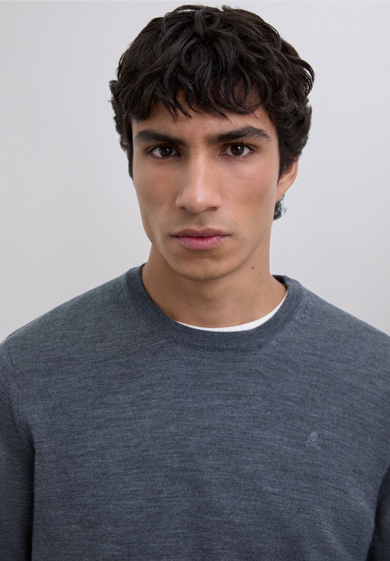 Grå stickad tröja med rund halsringning och texturerat mönster, med subtil logodetalj på bröstet. Närbild av plaggets tyg.