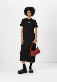 Juoda medvilninė T-shirt tipo suknelė su trumpomis rankovėmis, su logotipo detale. Modelis neša mažą raudoną rankinę ir dėvi juodas platformines batus bei kojines.
