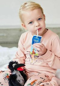 Litet barn med ljust hår som har på sig rosa pyjamas dricker ur en kartong med sugrör medan det håller i en svart och grå fågeltäcke.