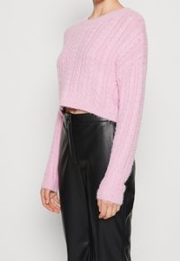 Suéter corto rosa hecho de un hilo suave y texturizado con un patrón de punto de cables y mangas largas, combinado con pantalones de cuero negros de talle alto.