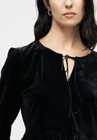 Black velvet dress features a round neckline, long puffed sleeves, and a tie closure at the neck. The skirt has a gathered texture at the hem.