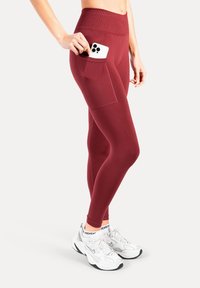 Leggings bordeaux avec une taille haute et une poche latérale contenant un smartphone blanc. Caractérisés par une texture lisse et extensible, associés à des chaussures de sport blanches.