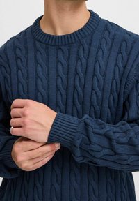 Pull en maille torsadée bleu marine avec un col rond et des poignets côtelés, présentant des motifs torsadés complexes et une texture douce.
