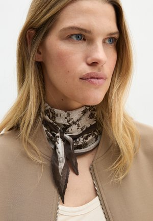 Blonde Frau mit blauen Augen, die eine beige Jacke und einen braun gemusterten Schal um den Hals trägt, leicht zur Seite blickend.