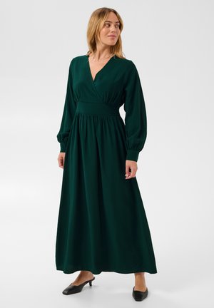 Femme portant une longue robe vert foncé à manches longues et des chaussures noires à talons bas, debout devant un fond clair uni.