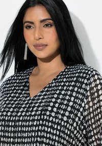 Sort og hvid top med V-hals, lavet af let stof med prikmønster. Langt, glat mørkt hår og hængende øreringe.