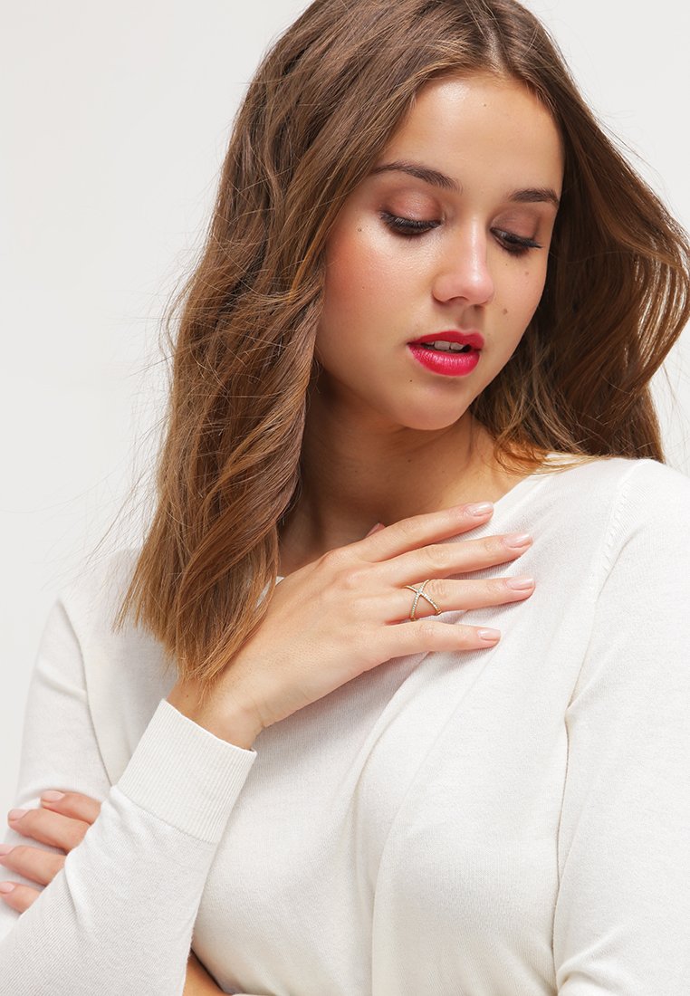 Gouden ring met een kruislings ontwerp, gedragen op een hand die op een witte trui rust. Lang, golvend bruin haar omlijst het gezicht. Rode lippen.