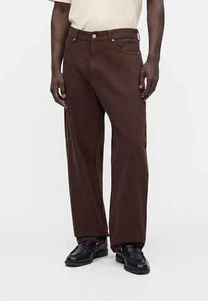 Homme portant un pantalon droit marron foncé, des chaussures habillées noires et une chemise beige clair à manches courtes, debout devant un fond blanc.