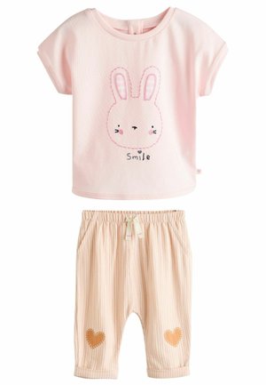 Camiseta de algodón rosa con un gráfico de un conejo y la palabra "Sonríe". Pantalones a rayas color beige con parches de corazones, cinturilla elástica y bolsillos.