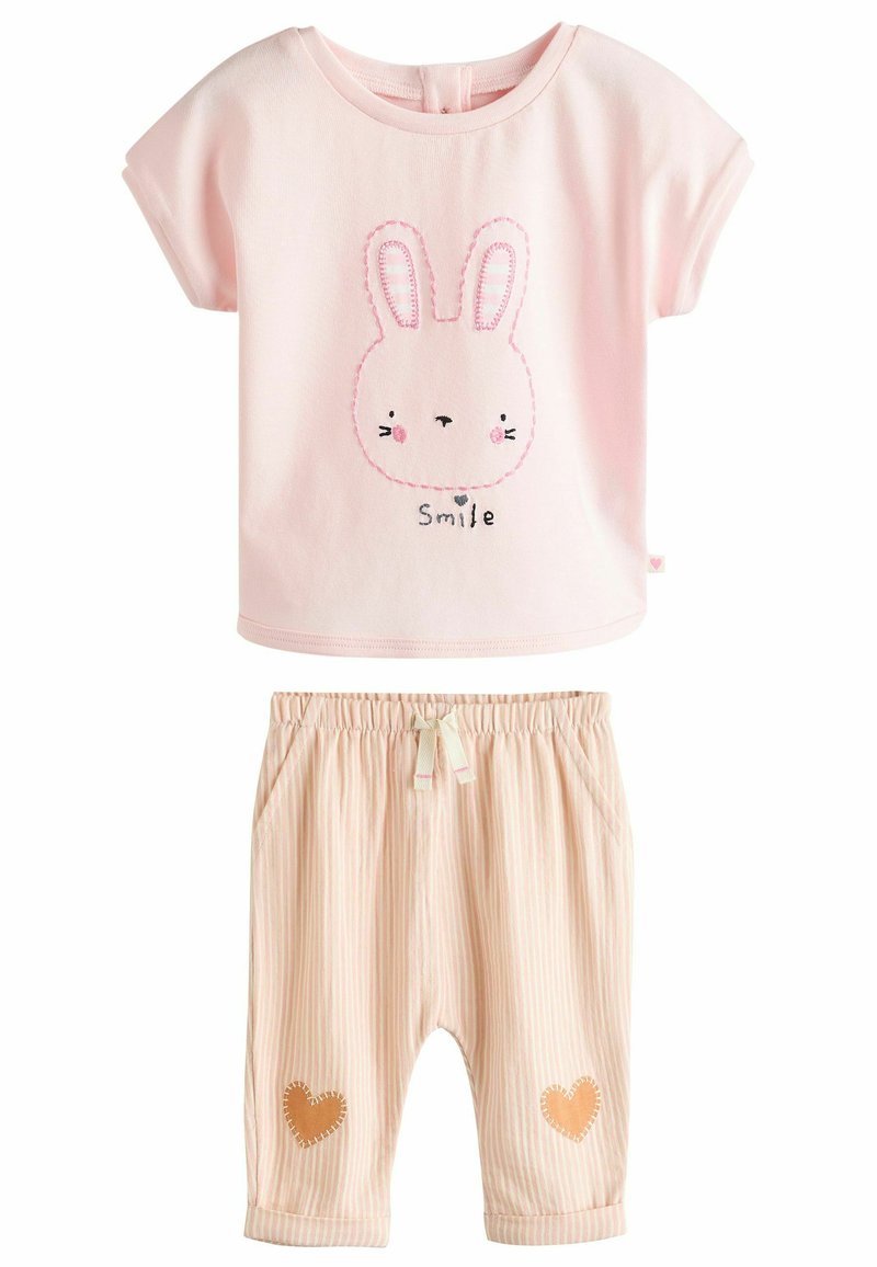Camiseta de algodón rosa con un gráfico de un conejo y la palabra "Sonríe". Pantalones a rayas color beige con parches de corazones, cinturilla elástica y bolsillos.
