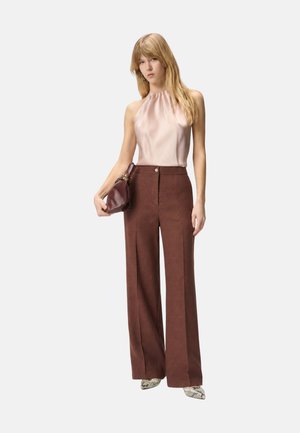 Donna che indossa un top in raso rosa blush senza maniche, pantaloni marroni a gamba larga e vita alta, che tiene una borsa in pelle marrone e indossa scarpe in pelle di serpente.