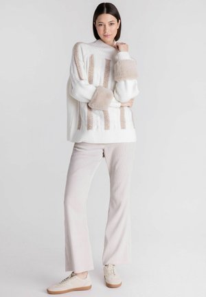 Vrouw staat, draagt een witte trui met beige strepen en bontboorden, lichtbeige ribfluwelen broek en beige sneakers tegen een effen achtergrond.
