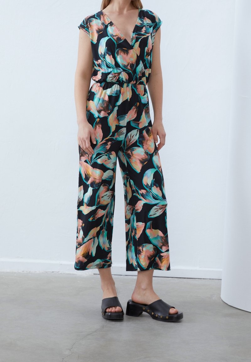 Femme portant une combinaison noire à fleurs avec des manches courtes et des jambes larges, associée à des sandales noires à talons sans bride.