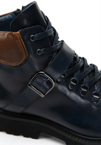 Botín de cuero azul oscuro con acentos de cuero marrón, que presenta una hebilla metálica, suela negra gruesa y cordones azules atravesados por ojales metálicos.