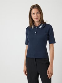 Femme portant un haut à rayures bleues marine à manches courtes avec un col volant, des rayures blanches et un pantalon noir, se tenant devant un fond uni.