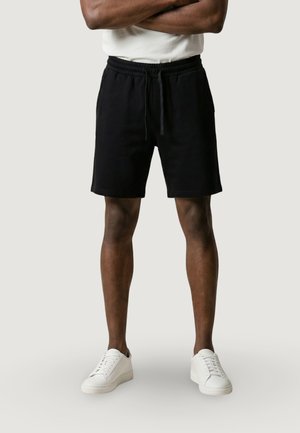 Mann trägt schwarze Shorts mit Kordelzug, weiße Sneakers und ein weißes Hemd, steht mit verschränkten Armen vor einem schlichten hellen Hintergrund.