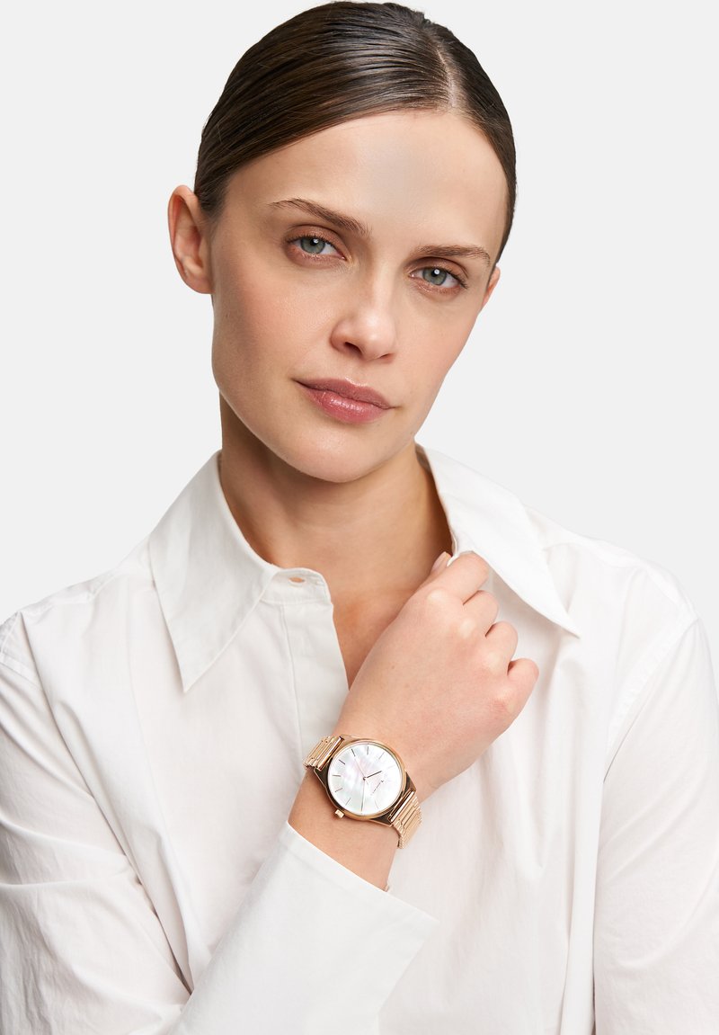 Gold-tone wristwatch with a round, iridescent mother-of-pearl dial, minimalistic markers, and a textured metal band, showcased on a wrist.