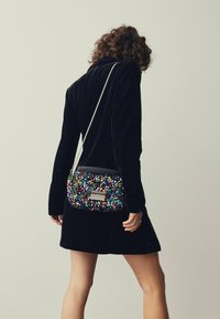 Svart sammetsskulderväska dekorerad med flerfärgade paljetter, med en silverkedja som axelrem och en rektangulär metallaccent på framsidan.