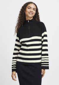 Pull-over rayé noir et blanc avec un col zippé mi-hauteur, présentant un tricot texturé. Manches longues avec un motif rayé s'étendant jusqu'aux poignets.