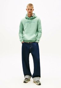 Myntrgrøn hoodie med frontlomme og logo, parret med mørkeblå vide bukser med grå manchet detaljer, båret med mønstrede sko.