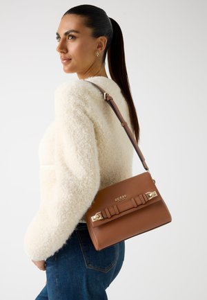 Borsa a spalla in pelle marrone con hardware dorato e dettaglio in nastro, abbinata a un maglione bianco peloso e jeans blu, su uno sfondo neutro.