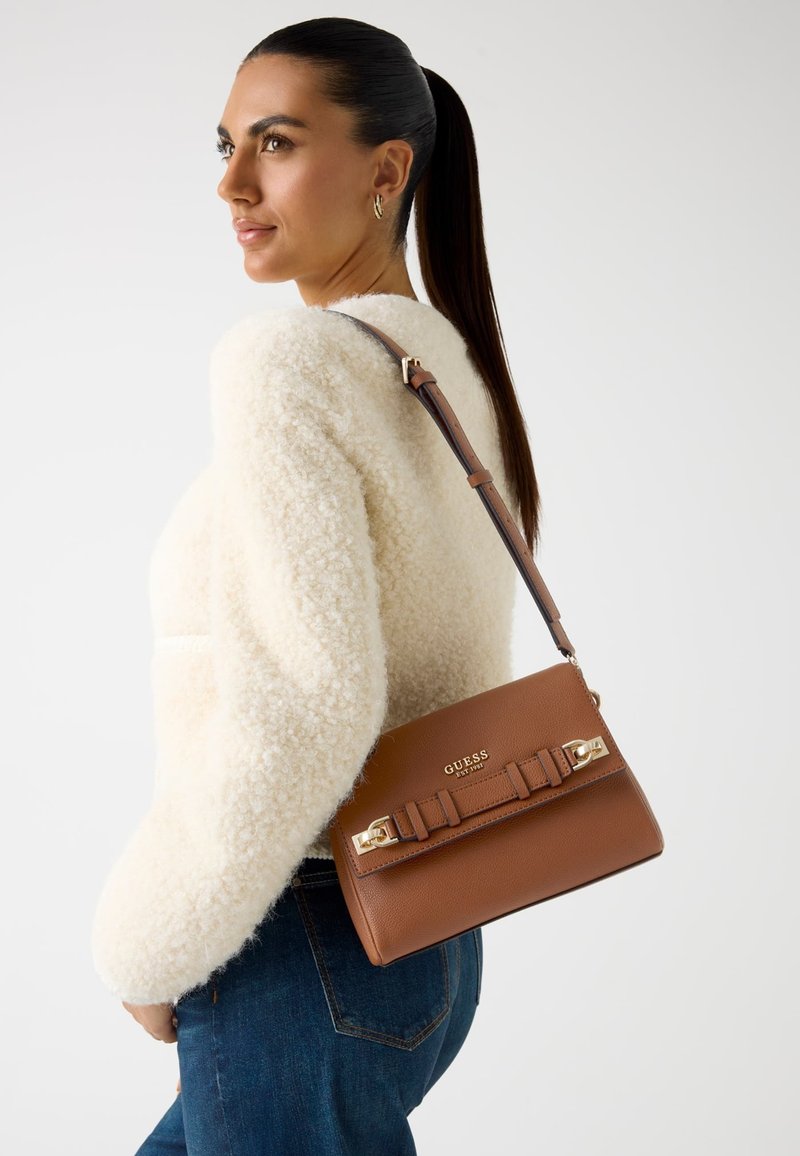 Braune Leder-Schultertasche mit goldenen Beschlägen und einem Bandakzent, getragen zusammen mit einem flauschigen weißen Pullover und blauen Jeans, vor einem neutralen Hintergrund.