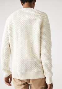 Sweter w kolorze kremowym wykonany na drutach, z teksturowanym wzorem w wstążki cukrowe, z prążkowanymi mankietami i pasem. Tył jest gładki, bez ozdób.