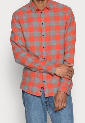 Mann trägt ein orange-grau kariertes Langarmhemd mit schwarzen Knöpfen und blaue Jeans, die Arme vor der Taille verschränkt.