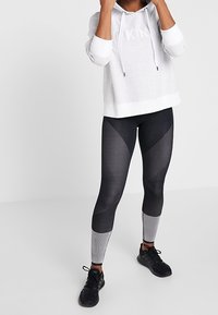 Grå hoodie med snoddar och frontlogotyp, kombinerad med svarta och grå mönstrade leggings med nätaccenter och en figurnära design. Svarta skor.