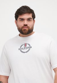 Baltas trumpas rankovėmis marškinėliai, pagaminti iš medvilnės, su centre esaniu "HILFIGER" logotipu, turinčiu tamsiai mėlynus ir raudonus akcentus bei laurų dizainu.