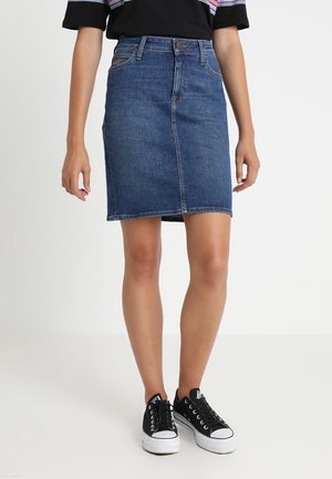 Vrouw draagt een blauwe denimrok tot op de knie, zwart-witte sneakers en een zwart T-shirt met gekleurde strepen op de mouwen.