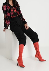 Blazer floreale nero, pantaloni larghi neri e stivali a stiletto rossi fino al ginocchio. Tessuto lucido e dettagli con vivace motivo floreale sul blazer.